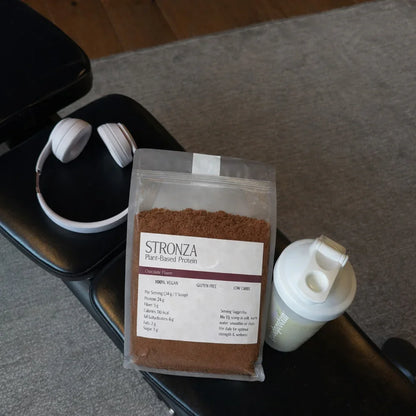 Stronza plant-based chocolate protein powder pouch with shaker bottle and headphones on gym bench